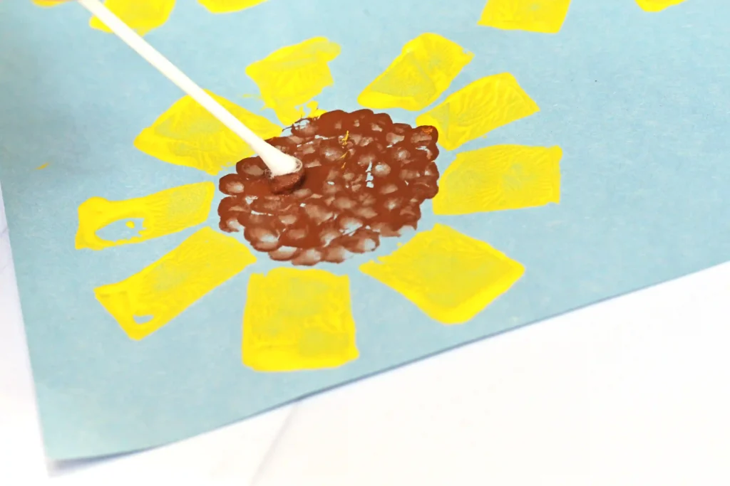 a cotton swab adding brown paint dots in the middle of a painted sunflower