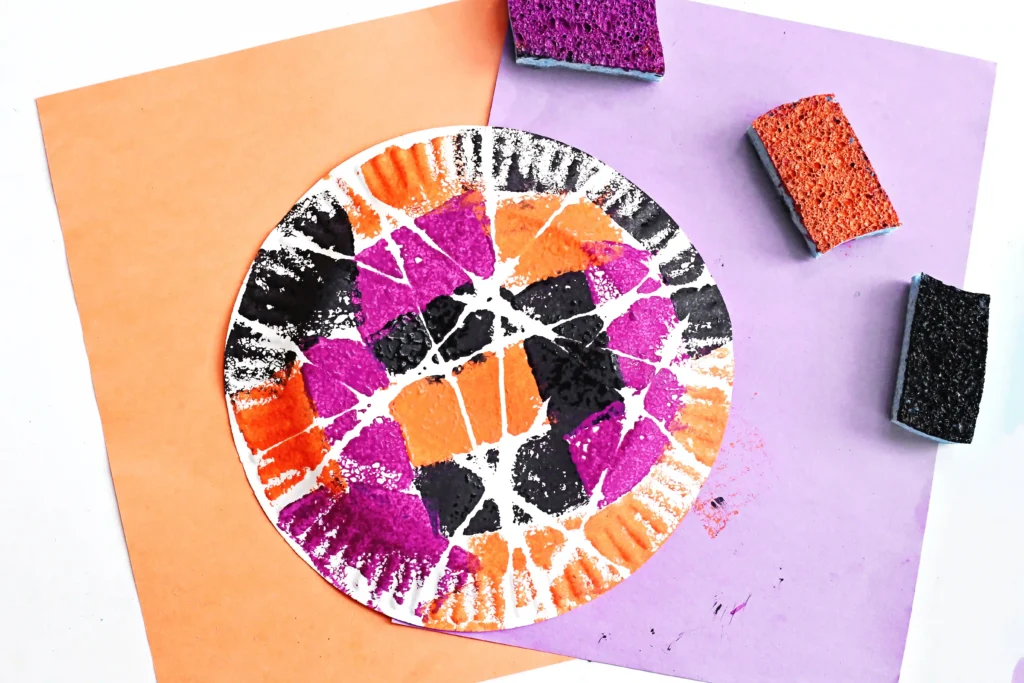 a Sponge Painted Spider Web on a paper plate with sponges and orange and purple paper in the background