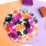 a Spider Web Painting on a paper plate with sponges and orange and purple paper in the background