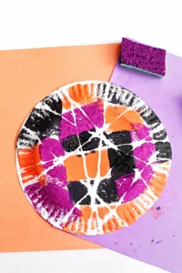 a Spider Web Painting on a paper plate with sponges and orange and purple paper in the background