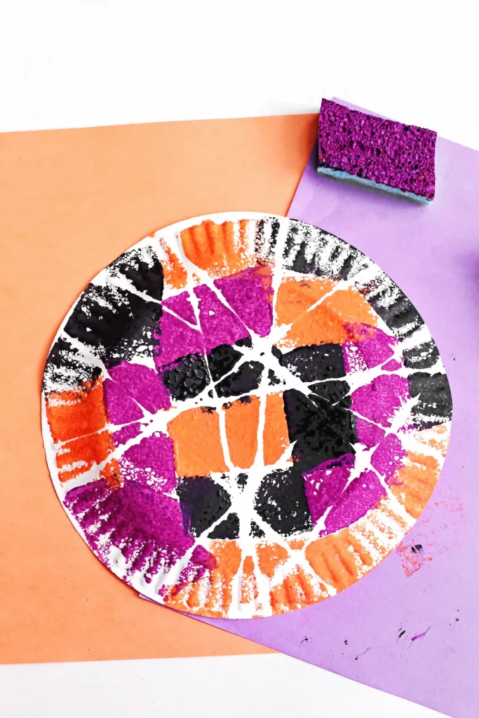 a Spider Web Painting on a paper plate with sponges and orange and purple paper in the background