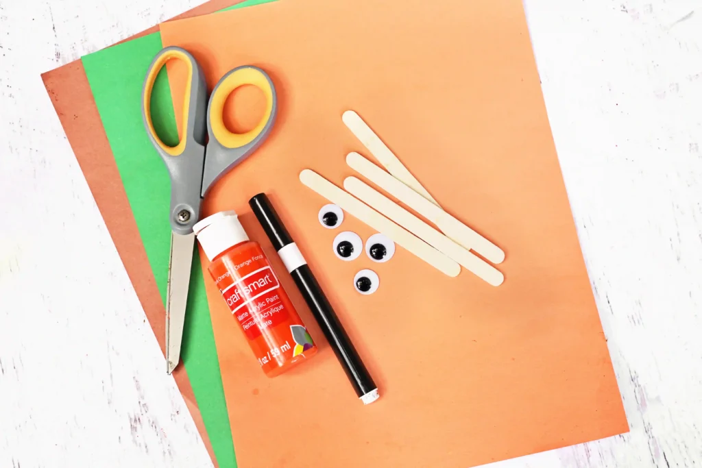 supplies for this fall book craft- brown, green, and orange construction paper, scissors, popsicle sticks, googly eyes, a black marker, and orange paint