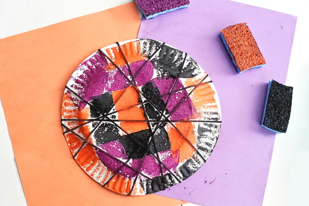 a Spider Web Painting on a paper plate wrapped with black yarn with sponges and orange and purple paper in the background