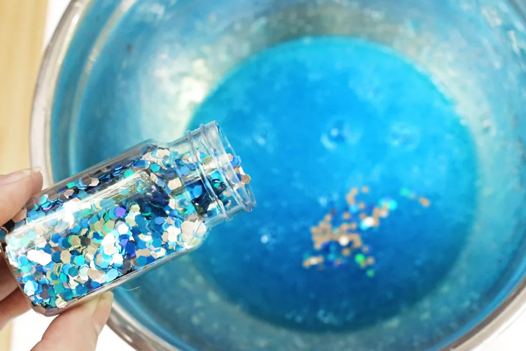 blue and silver glitter being added to a bowl of blue slime