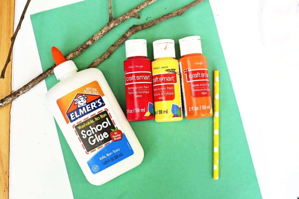 supplies for the campfire painting: sticks, glue, paint brush, straw, green paper, and red, yellow, and orange paint