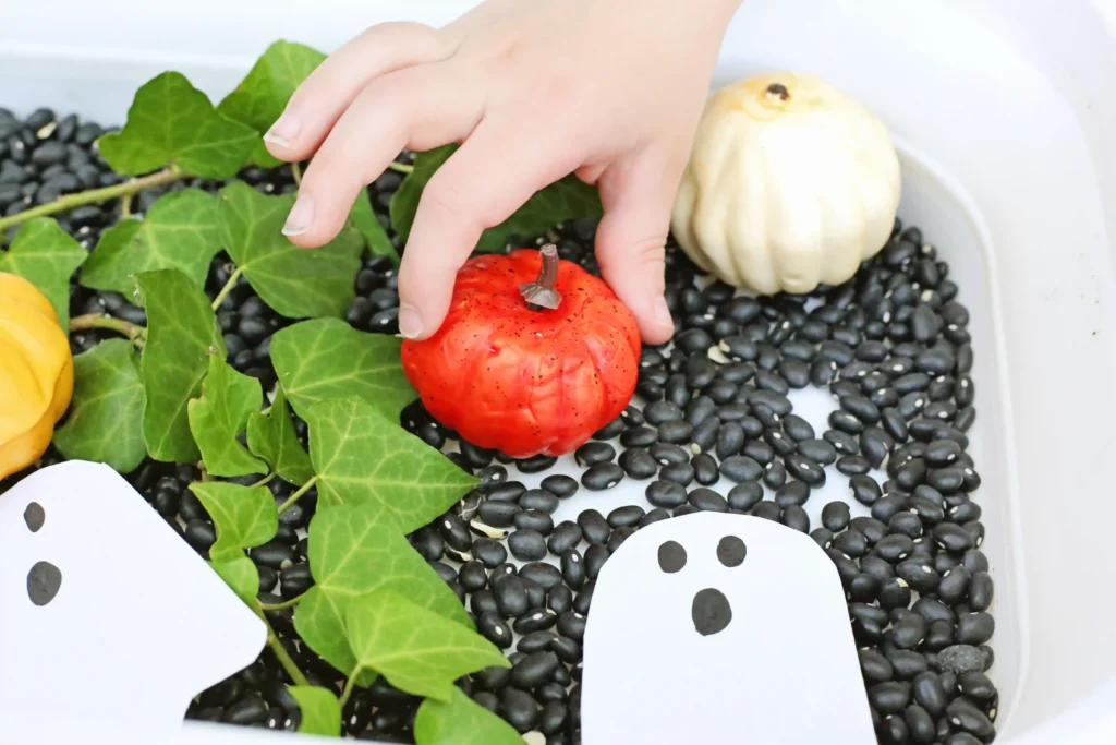 a kid picking up a fake pumpkin for a bin with leaves and black beans