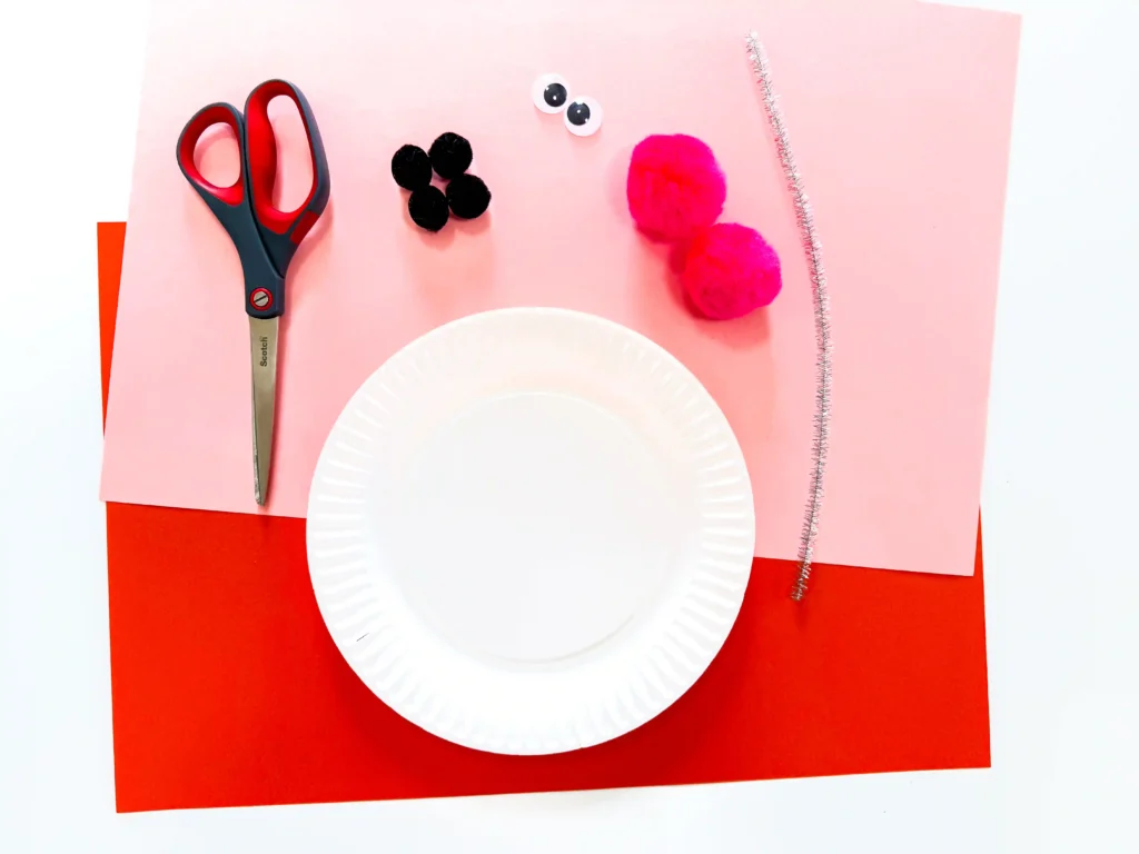 supplies for the snowman paper plate craft- orange and pink construction paper, a paper plate, black pom poms, pink pom poms, googly eyes, and scissors.