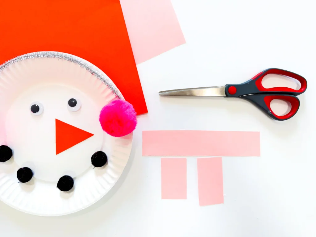 a paper plate decorated to look like a snowman next to pink rectangular pieces of construction paper