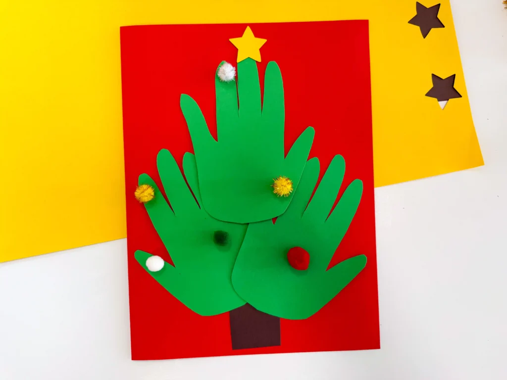 red construction paper with a brown square and green handprints, a star, and pom poms