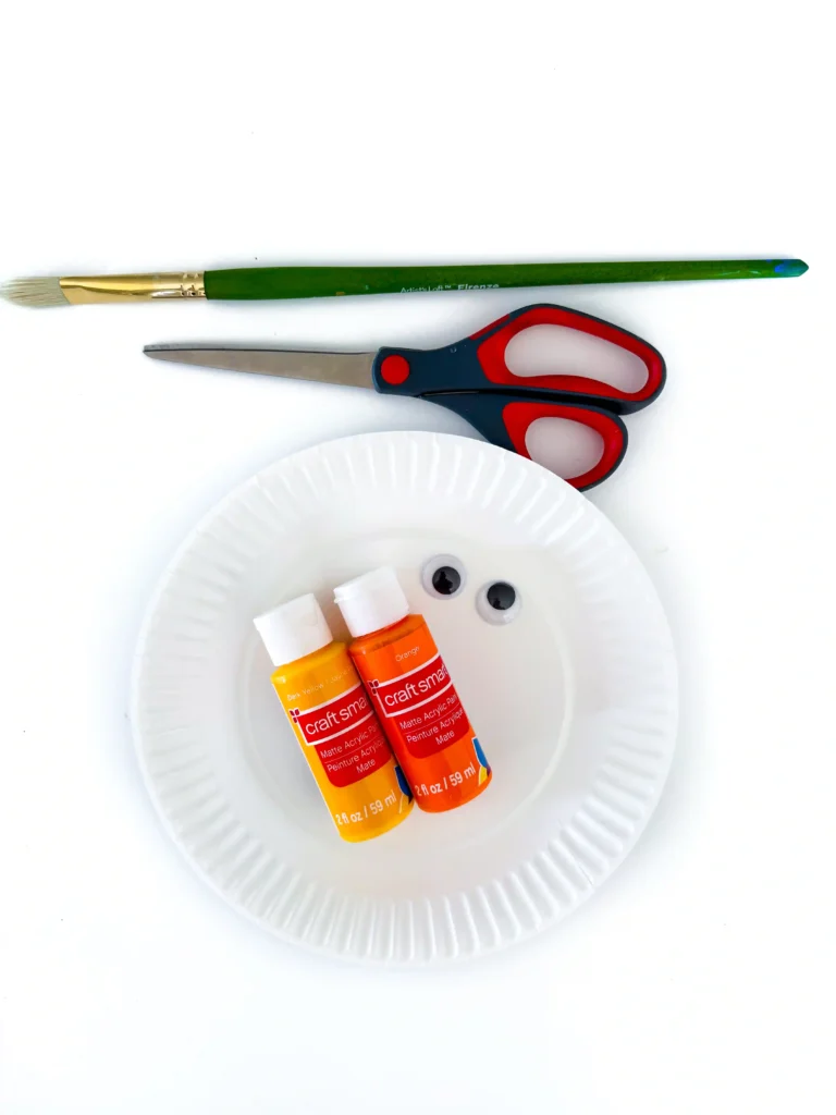 supplies for the paper plate candy corn craft: a paper plate, googly eyes, scissors, paint brush, and yellow and orange paint