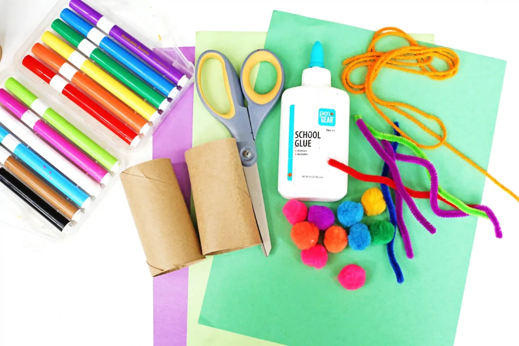 supplies for the monster binoculars: markers, colored construction paper, scissors, glue, toilet paper rolls, pom poms, pipe cleaners, and string