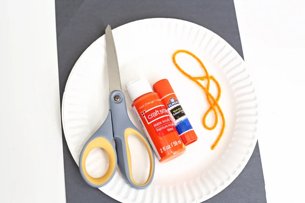 supplies for the jack-o-lantern craft: paper plate, scissors, orange paint, glue, orange string, and black construction paper