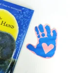 a blue handprint with a heart in the middle on pink paper next to a book