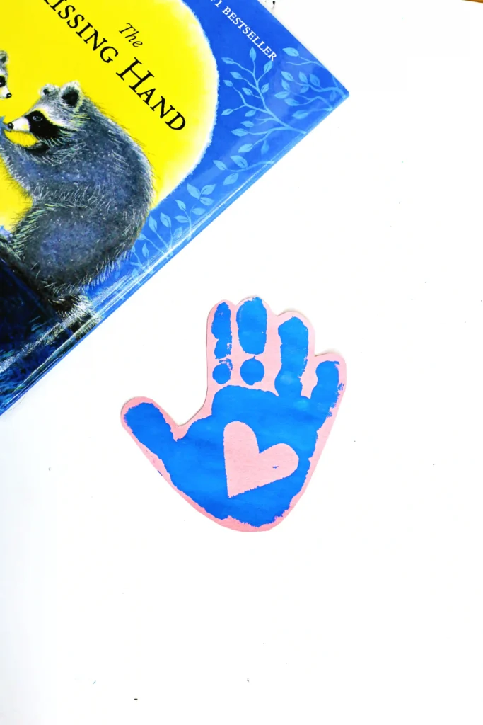 a blue handprint with a heart in the middle on pink paper next to a book