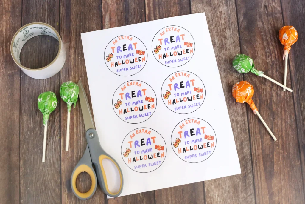 supplies for the Printable Halloween tags: scissors, tape, lollipops, and tags that read "an extra treat to make halloween super sweet"