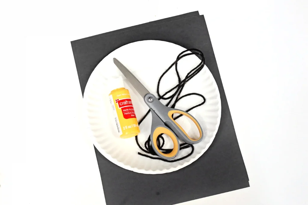 supplies for the bat mobile paper plate craft: black paper, a paper plate, yellow paint, string, scissors, and glue