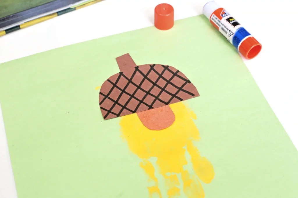 a yellow handprint with an acorn cap and orange nose on green construction paper
