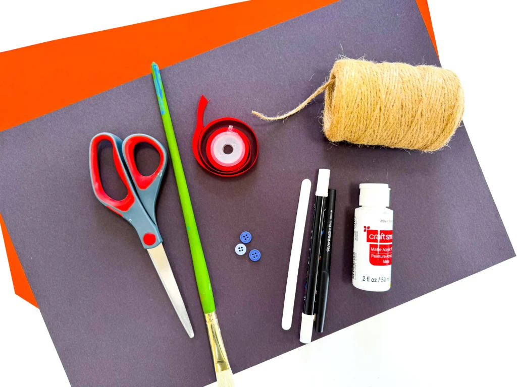 supplies for the snowman popsicle stick ornament: black and orange paper, scissors, paintbrush, ribbon, twine, white paint, buttons, a popsicle stick and a black and white marker