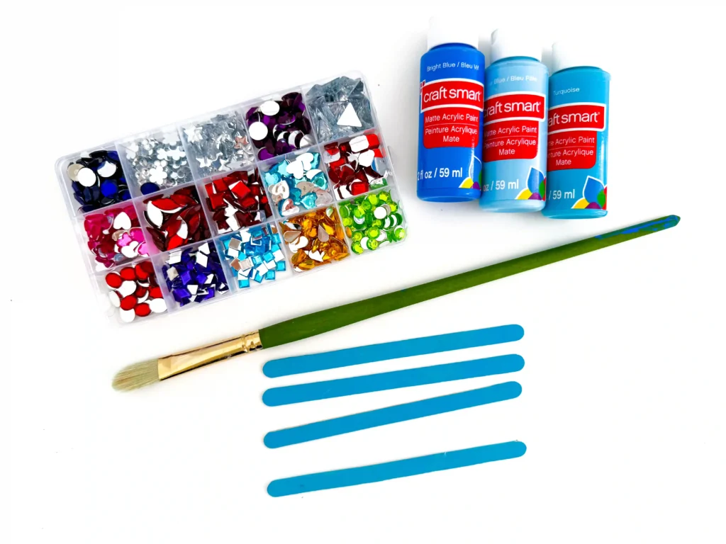 supplies for the popsicle stick snowflake craft: blue paint, paintbrush, popsicle sticks, and gems