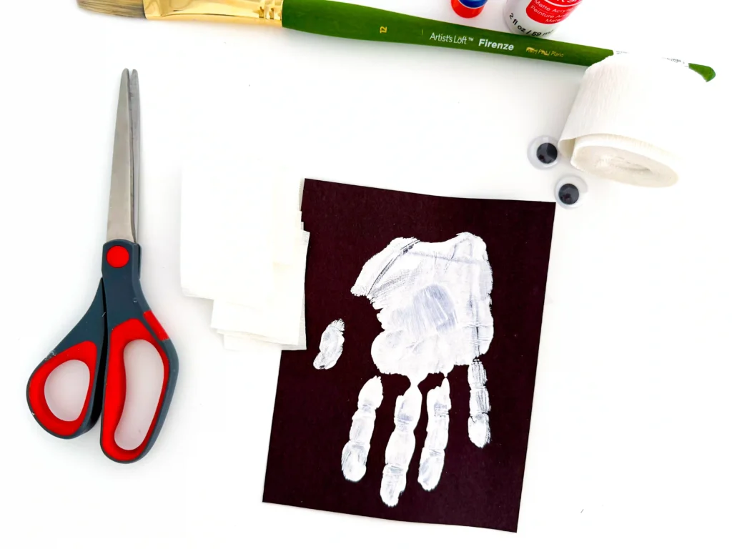 a white handprint on black paper next to white streamer and scissors