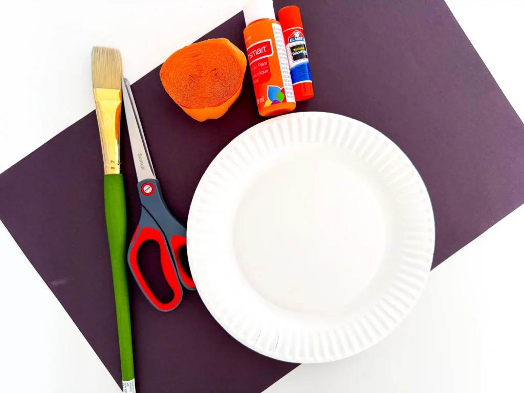 supplies for the pumpkin paper plate craft: a paper plate, orange paint, paintbrush, scissors, glue, black construction paper, and orange streamer