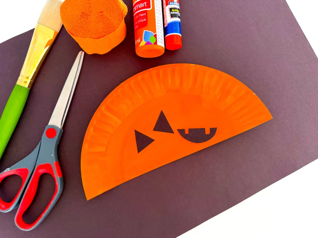 half of a paper plate painted orange and a jack-o-lantern face cut out of black construction paper