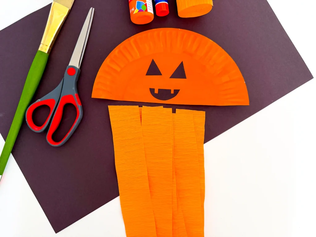 half of a paper plate painted orange with a jack o lantern face next to strips of orange streamer