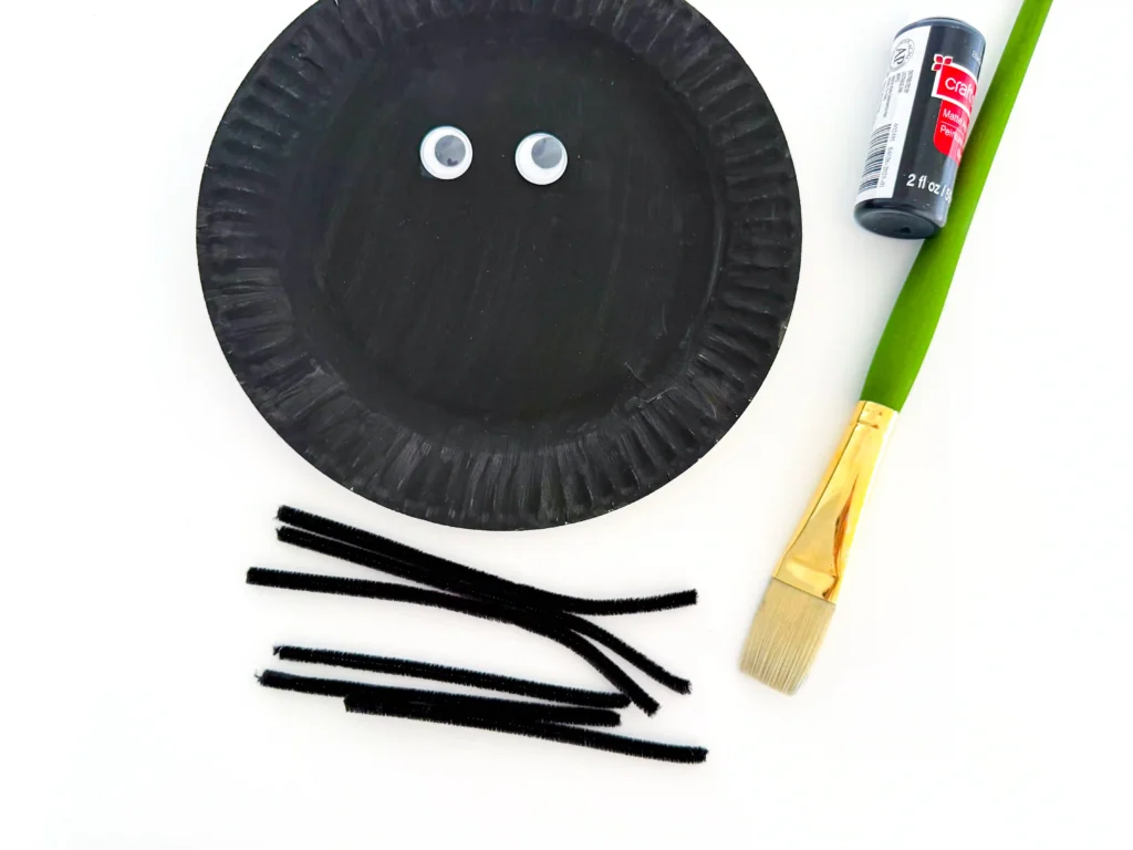 googly eyes glued on a plate painted black next to small black pipe cleaners