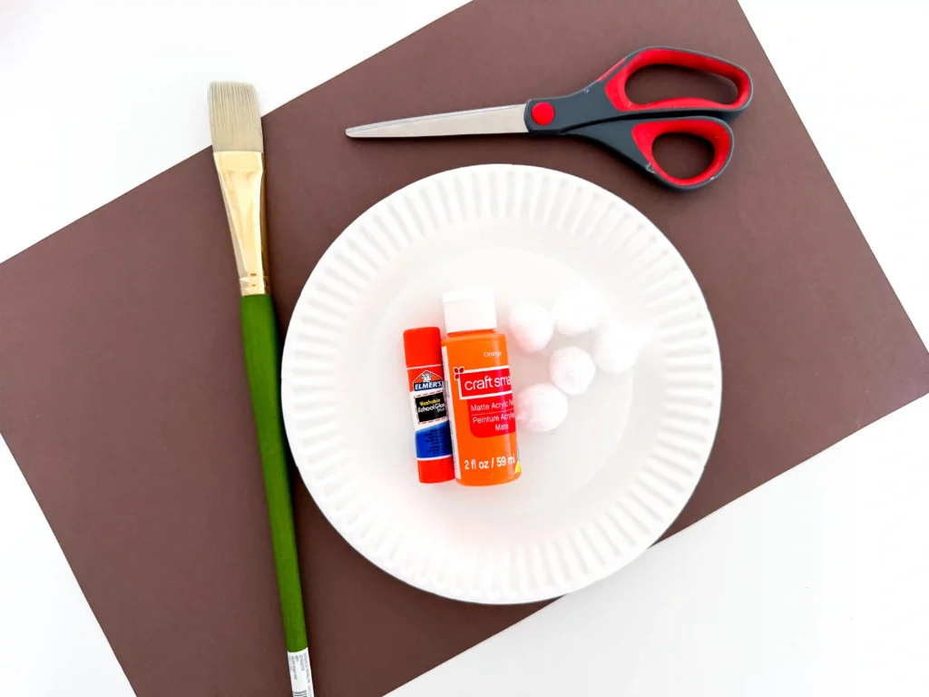 supplies for the pumpkin paper plate craft- paint brush, brown construction paper, glue, orange paint, white pom poms, and scissors