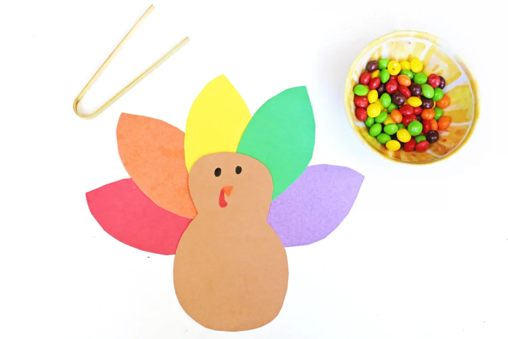 a turkey made out of colored construction paper, a bowl of skittles, and tweezers