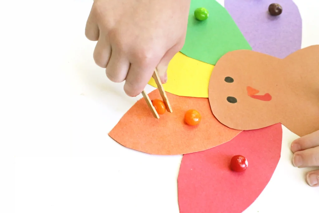 a kid using tweezers to put skittles on construction paper turkey feathers